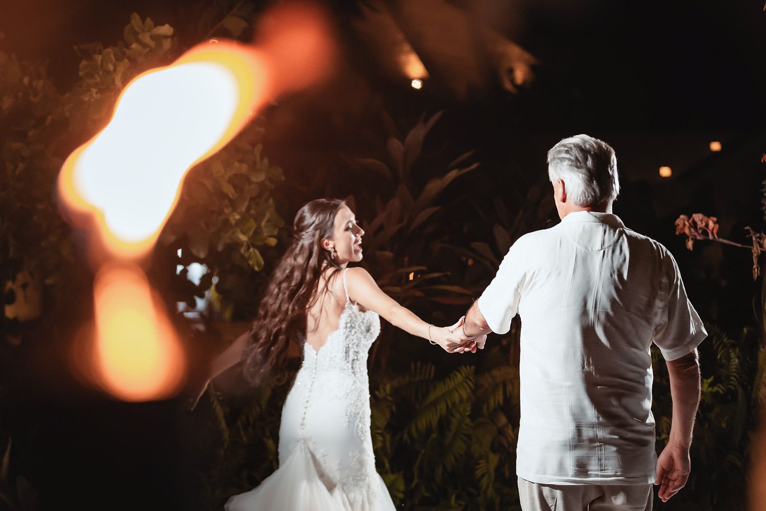 Bride and groom hold hands by a glowing fire torch during an outdoor nighttime wedding celebration.