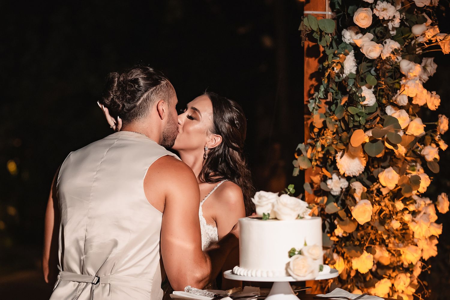 Bride and groom sharing a kiss beside a white wedding cake adorned with flowers at an outdoor evening reception.