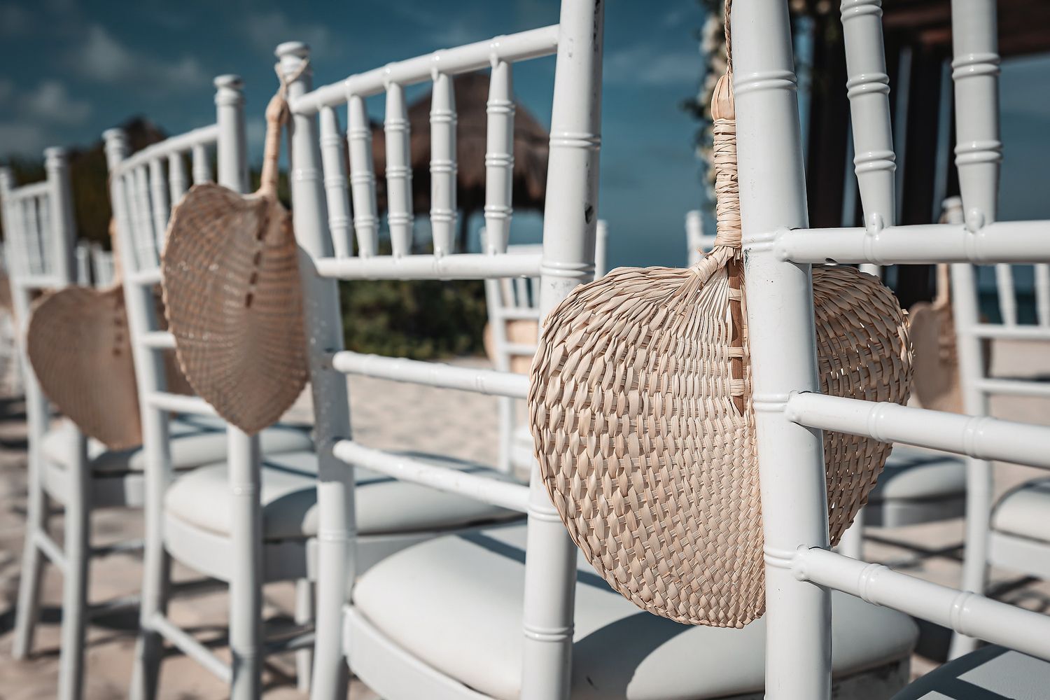 White chiavari chairs with woven fan decorations hanging on the backs, set up for an outdoor wedding ceremony.