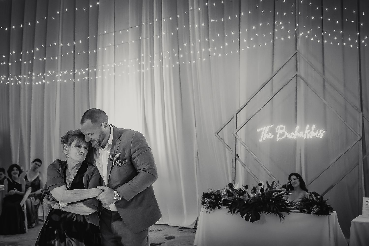 Black and white wedding reception photo of groom sharing emotional dance with mother near glowing neon sign.