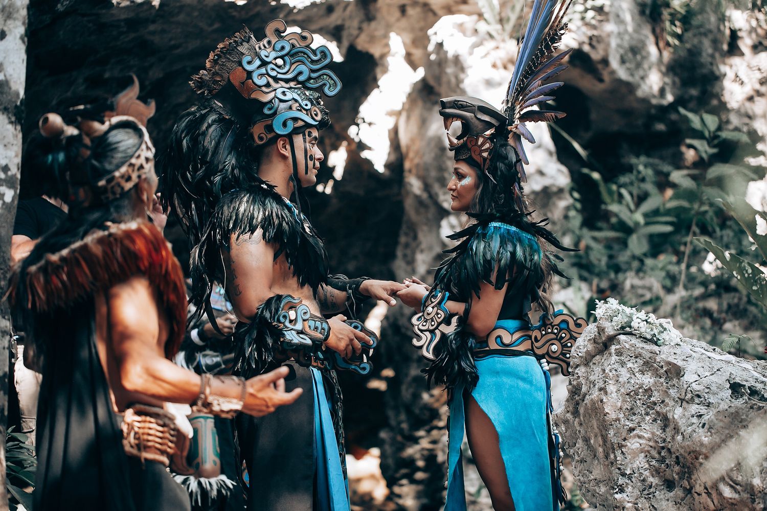 Three performers in detailed Mayan warrior costumes conversing near rocky cave in jungle setting.