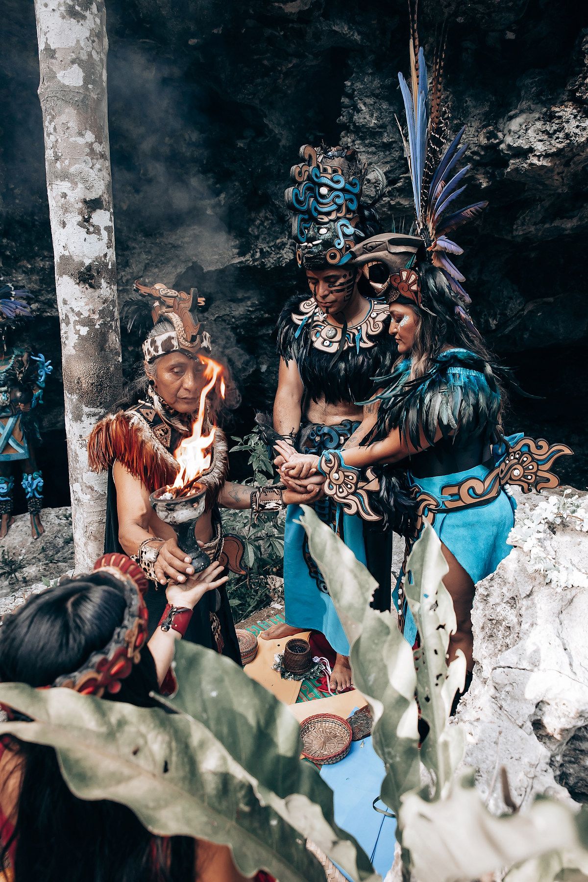 Mayan ceremonial performers in feathered headdresses conduct a fire ritual with traditional offerings in a cave setting.