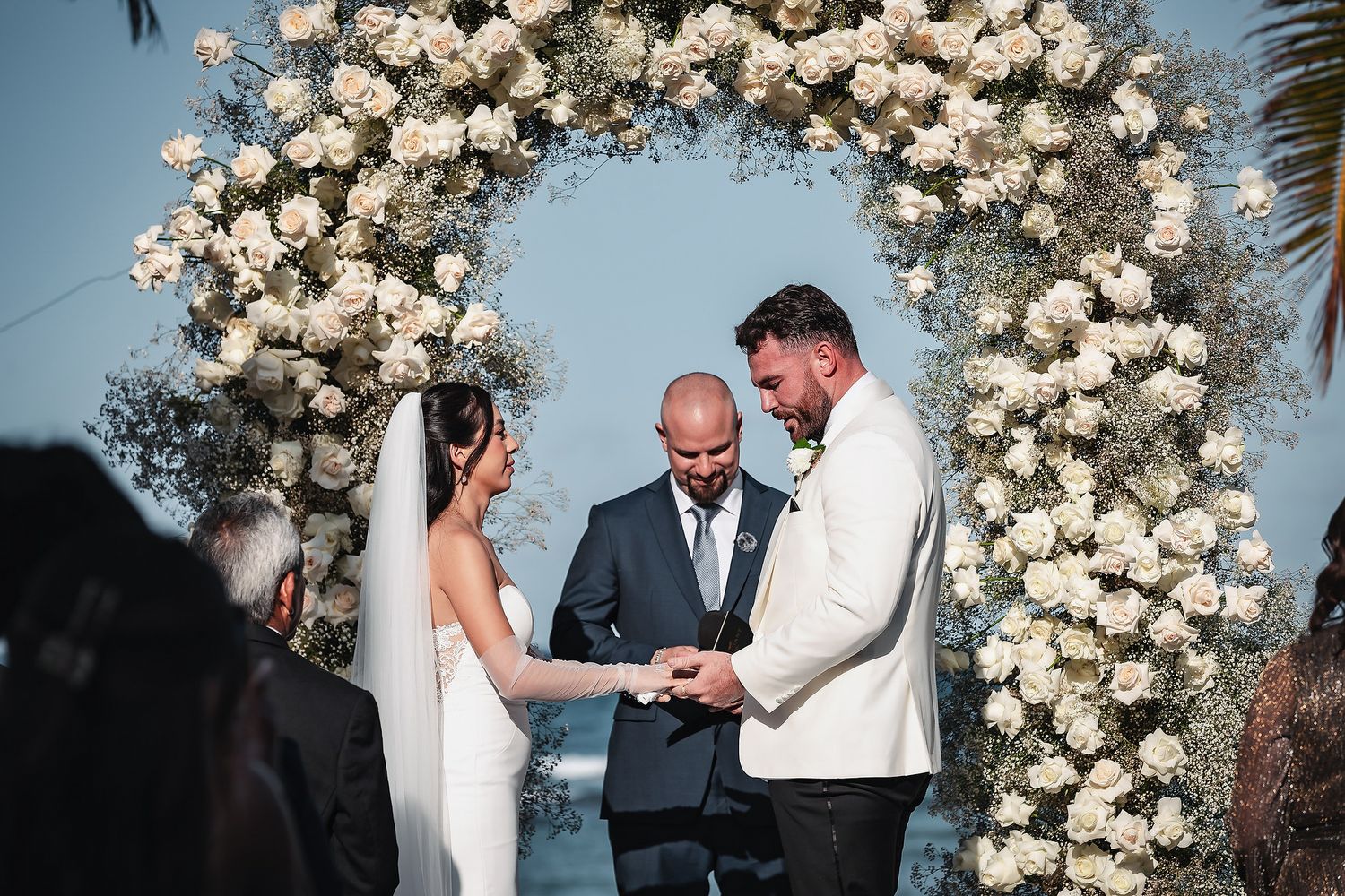 Couple exchanges vows under a stunning white floral arch at an outdoor wedding ceremony by the ocean.