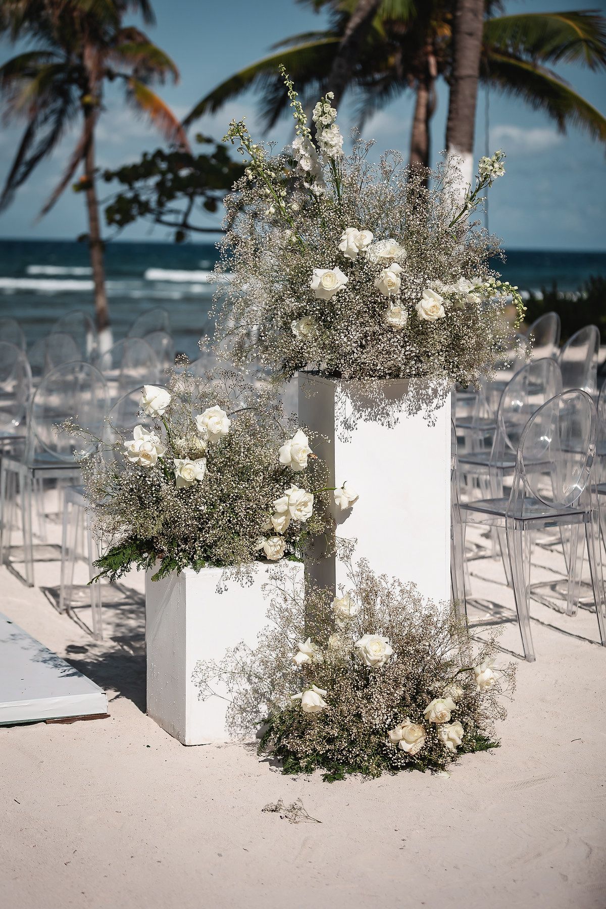 Beach wedding ceremony setup with white floral arrangements featuring roses and baby's breath on white pedestals, with clear chairs on sand.