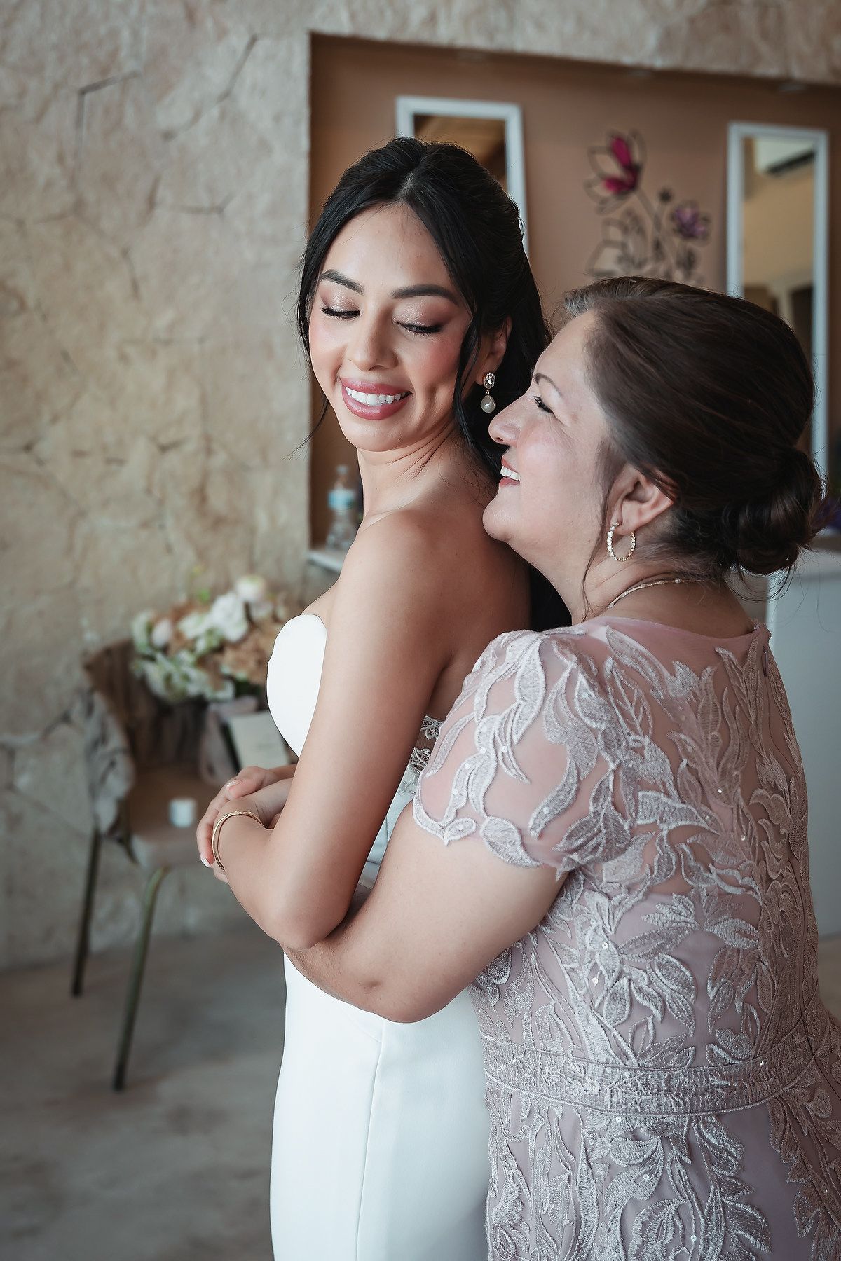 A bride in a white gown smiles warmly as an older woman in lace embraces her during wedding preparations.