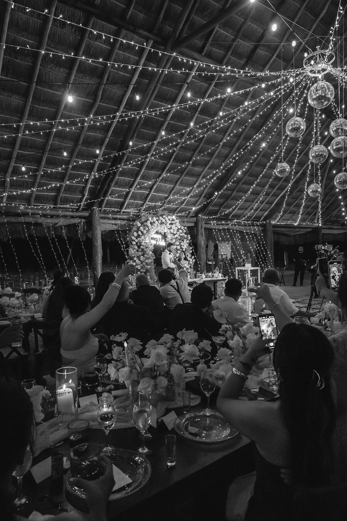 Black and white wedding reception with string lights and disco balls as guests raise glasses in a toast.