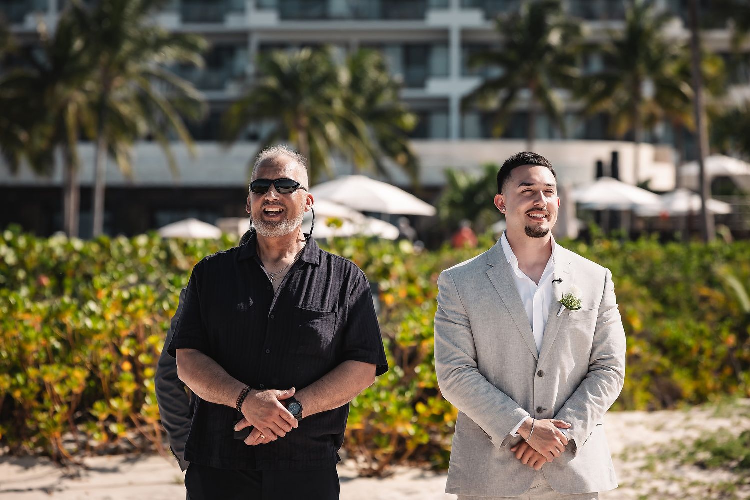 Two men stand on a beach near a resort, one in a black shirt and sunglasses, the other in a light gray wedding suit.