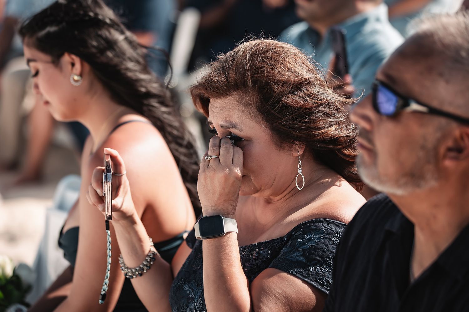 Emotional woman wiping tears at outdoor event, wearing dark dress and smartwatch, with blurred guests in background.