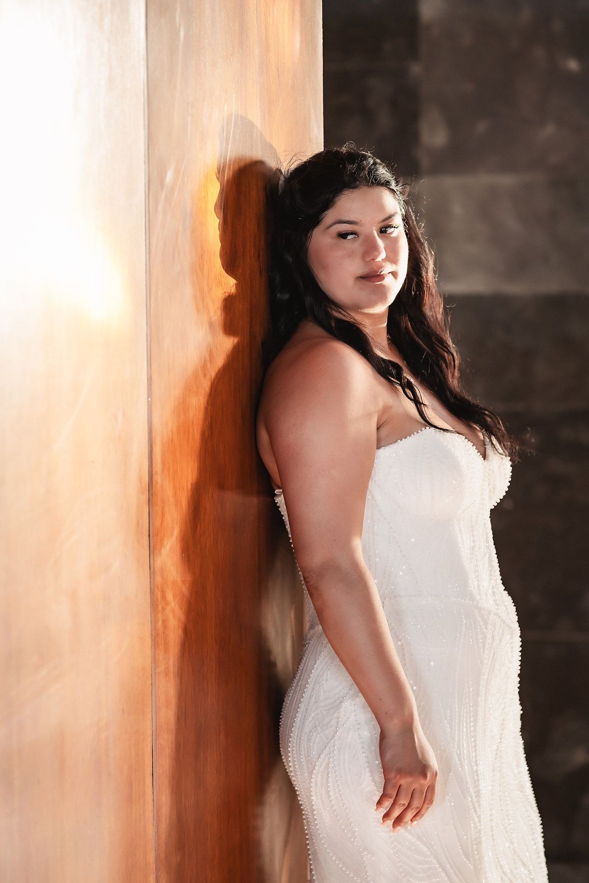 A woman in a white strapless wedding gown leans against a warm-lit wooden column in an elegant venue.