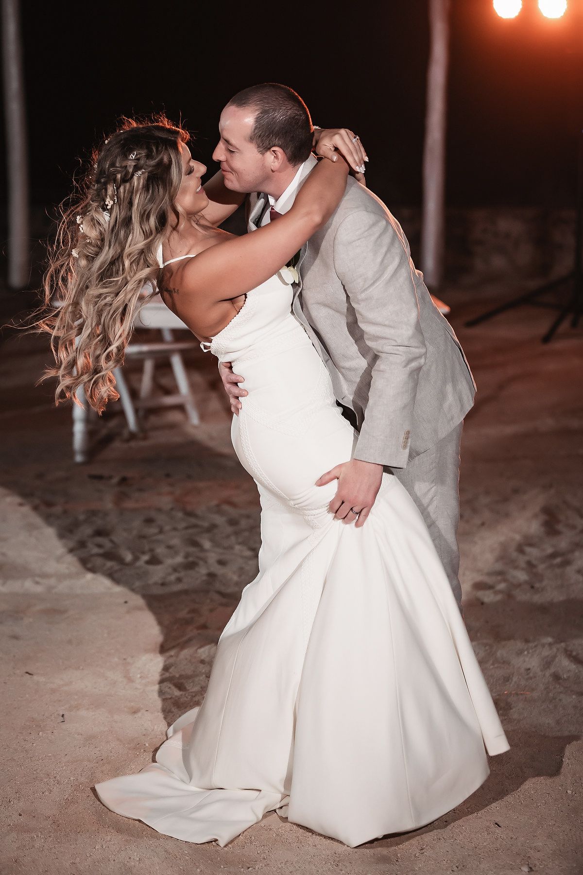 Bride and groom share a passionate dip kiss on sandy beach during their evening wedding celebration.