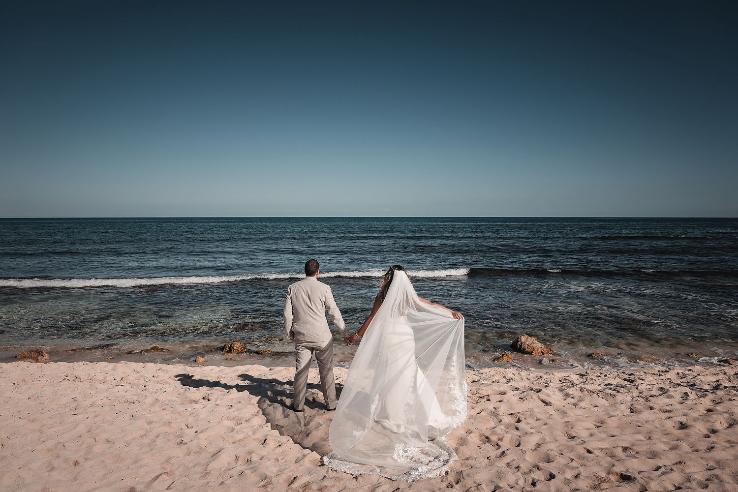 Bride and groom standing on sandy beach facing ocean, wedding veil flowing in wind, romantic seaside ceremony photo.
