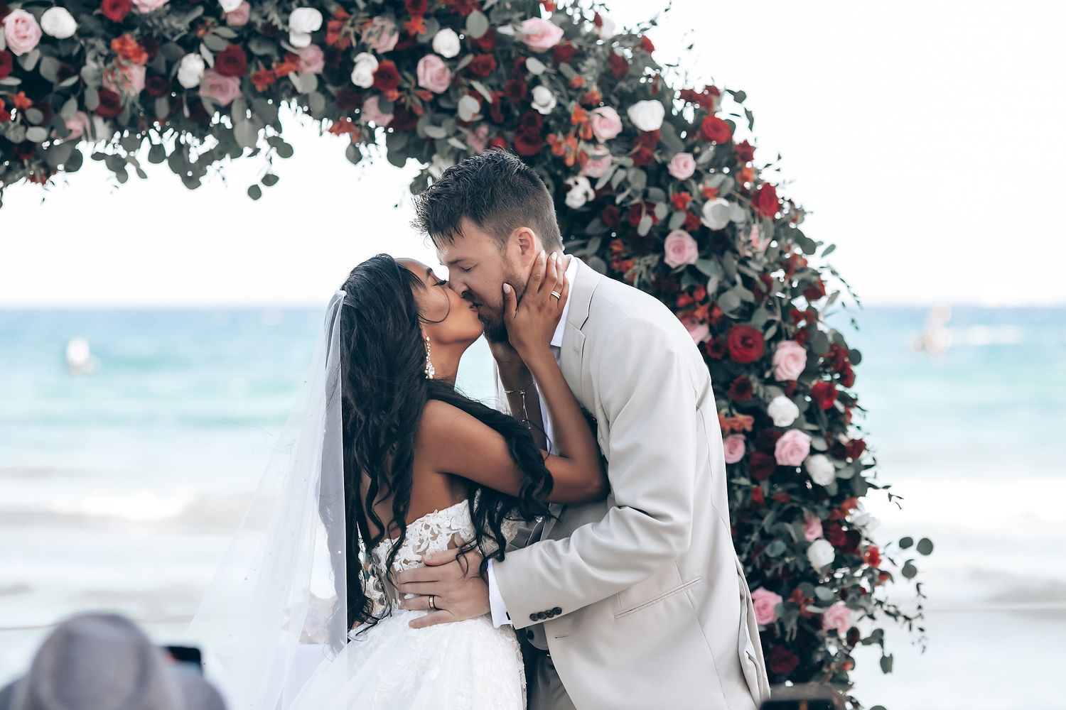 Romantic beach wedding ceremony with couple embracing under floral arch by ocean at sunset.