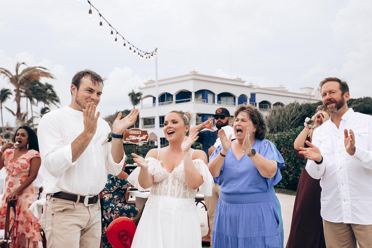 Wedding party and guests celebrate outdoors with raised hands and joyful expressions.