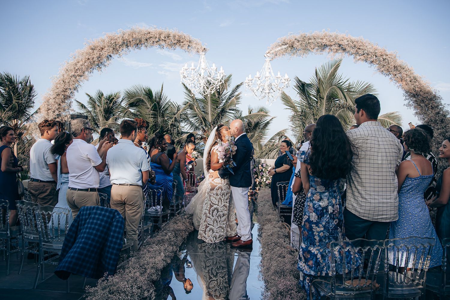 A rustic outdoor wedding ceremony takes place under a curved wooden arch decorated with white flowers and greenery.
