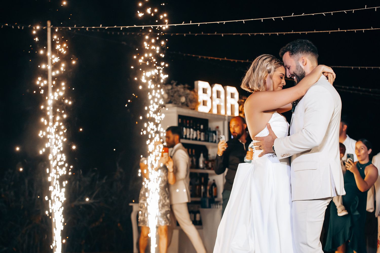 Newlyweds dancing with sparklers creating magical light effects at night reception.