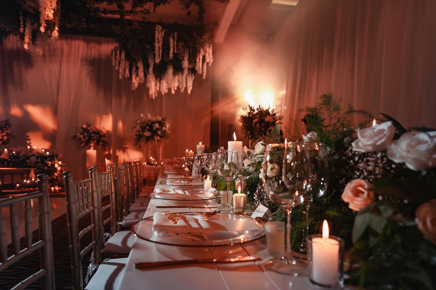 Elegant wedding reception table setting with candles, crystal chandeliers, and floral arrangements in warm romantic lighting.