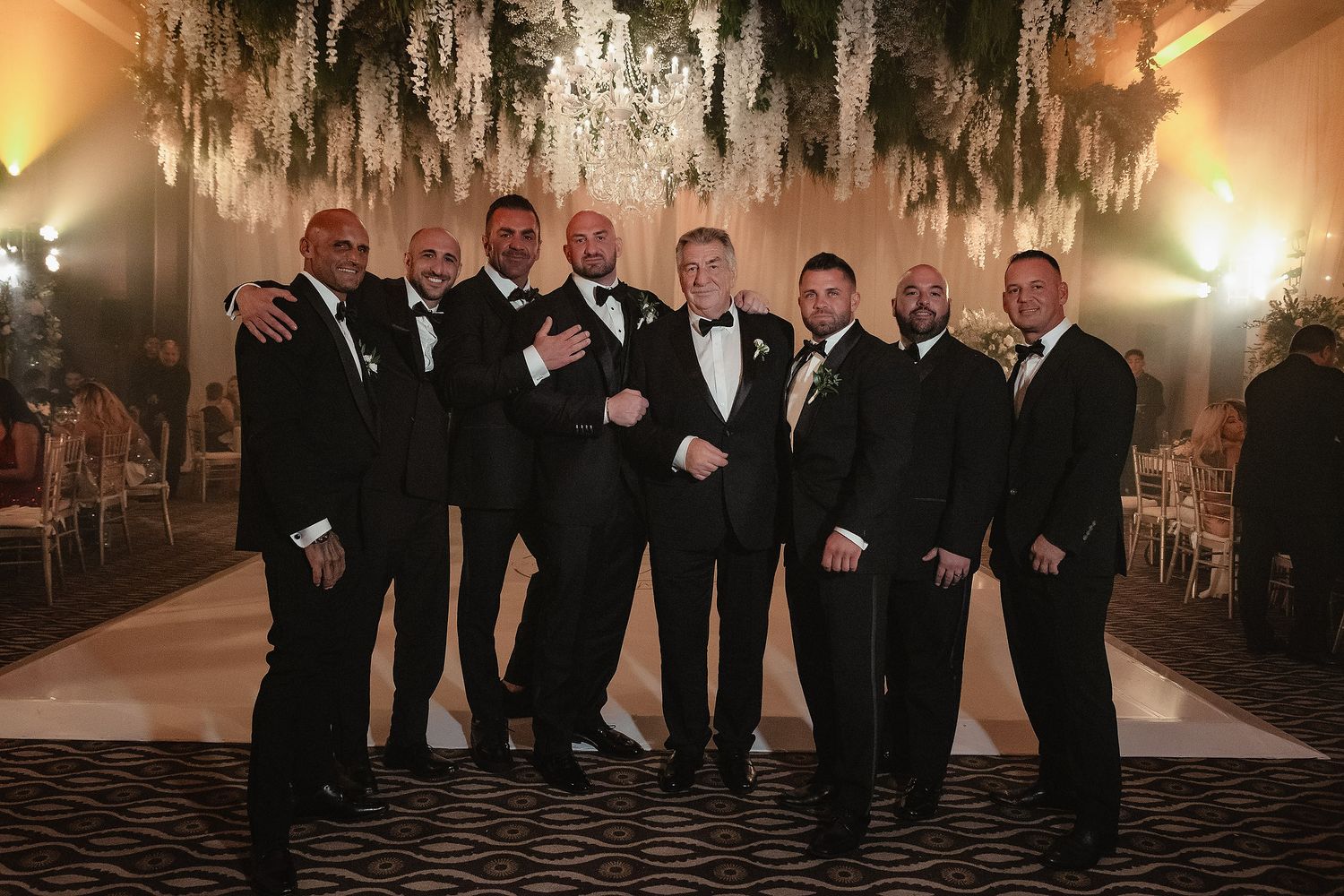 A group of groomsmen in black tuxedos pose together under elegant hanging decorations at a wedding reception.