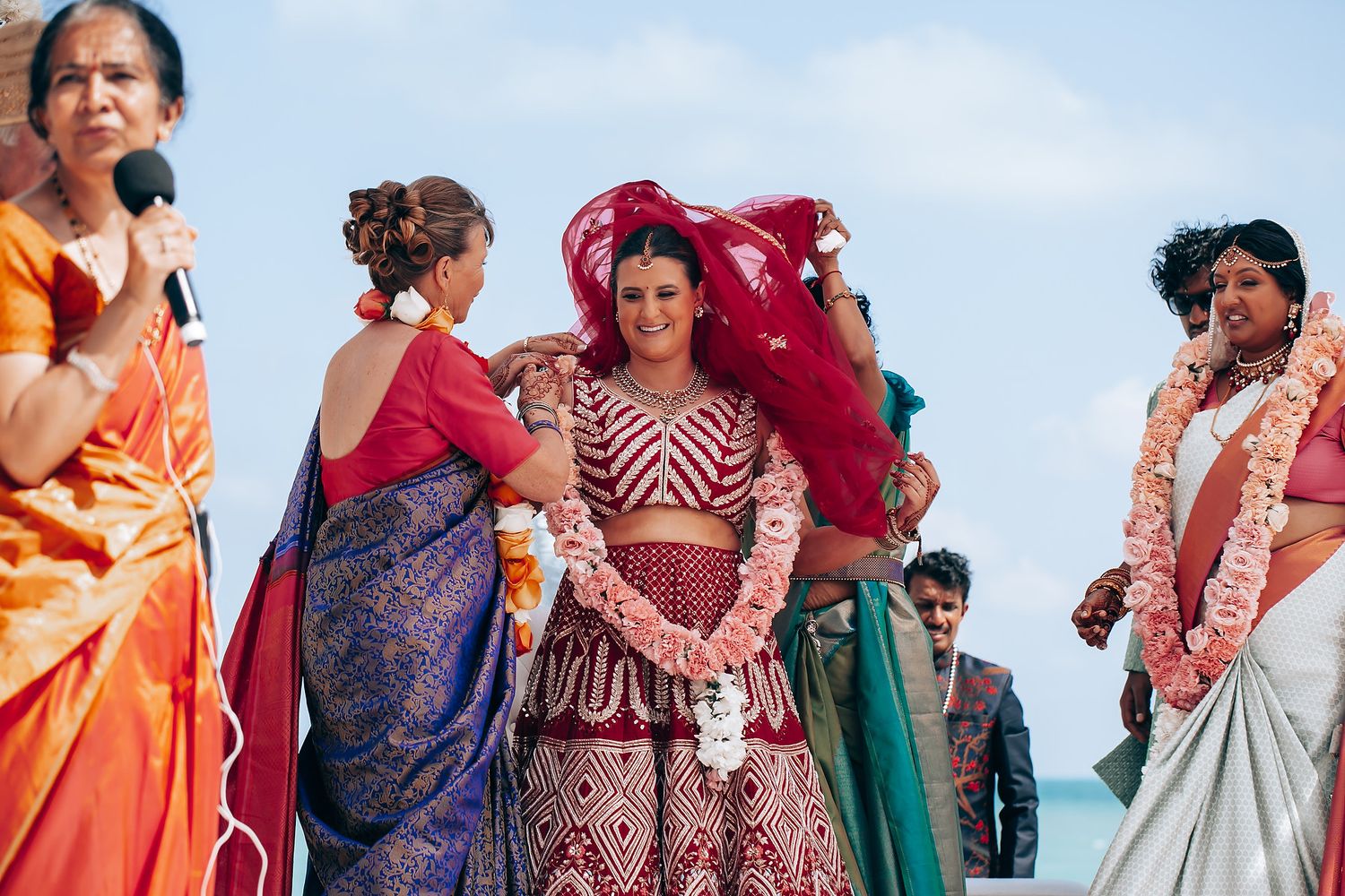 Guests in colorful traditional Indian attire gather at a beachside wedding ceremony under floral decorations.