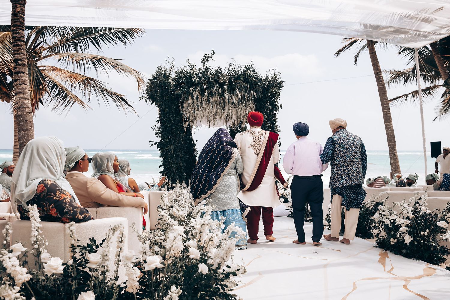 Guests gather at an outdoor wedding ceremony under palm trees with white decor and floral arrangements.