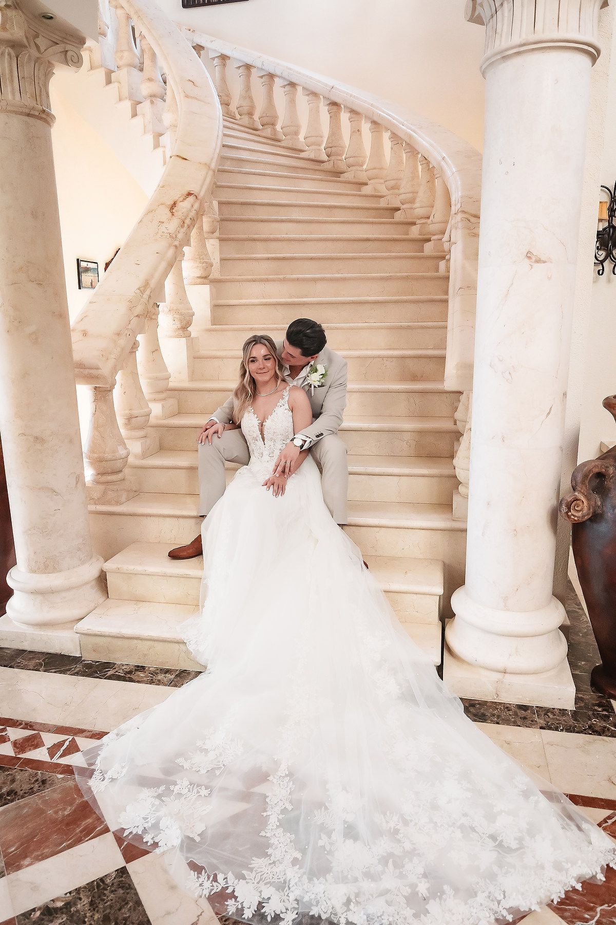A wedding couple poses on an elegant white marble staircase with classical columns.