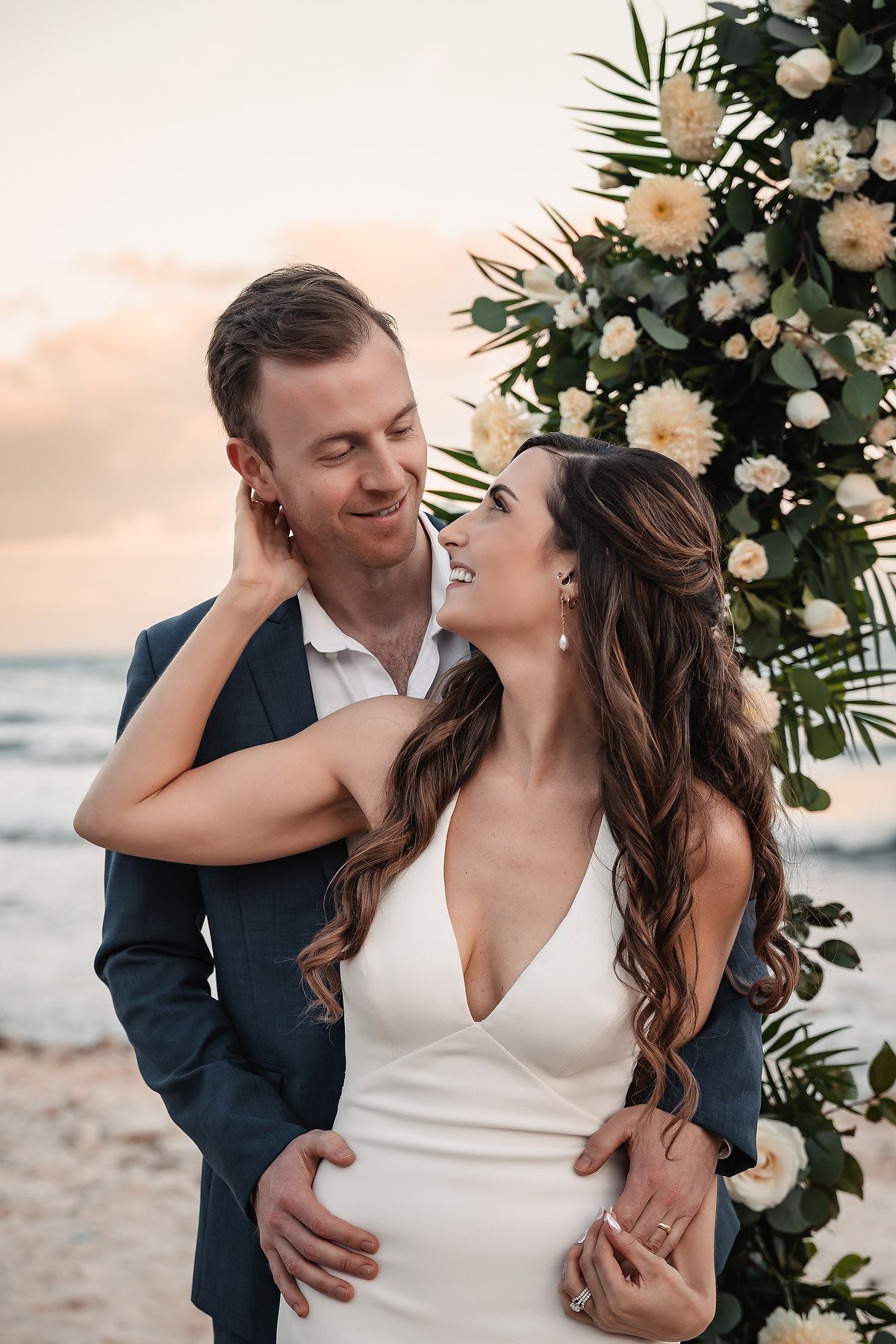 Romantic beach wedding portrait with floral arch and sunset backdrop.