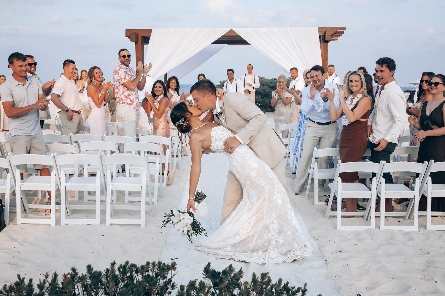 Wedding ceremony under a white canopy with guests seated on white chairs by the ocean.