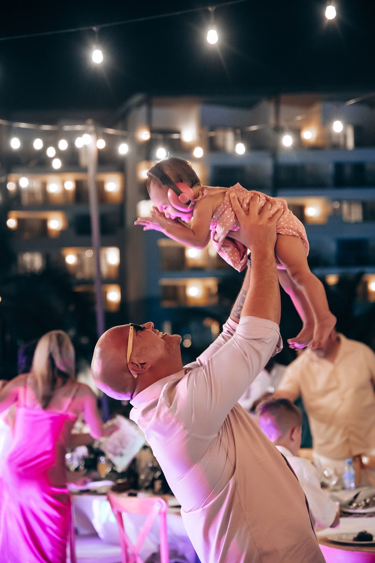 People in white attire dancing and celebrating at an evening party with pink lighting.
