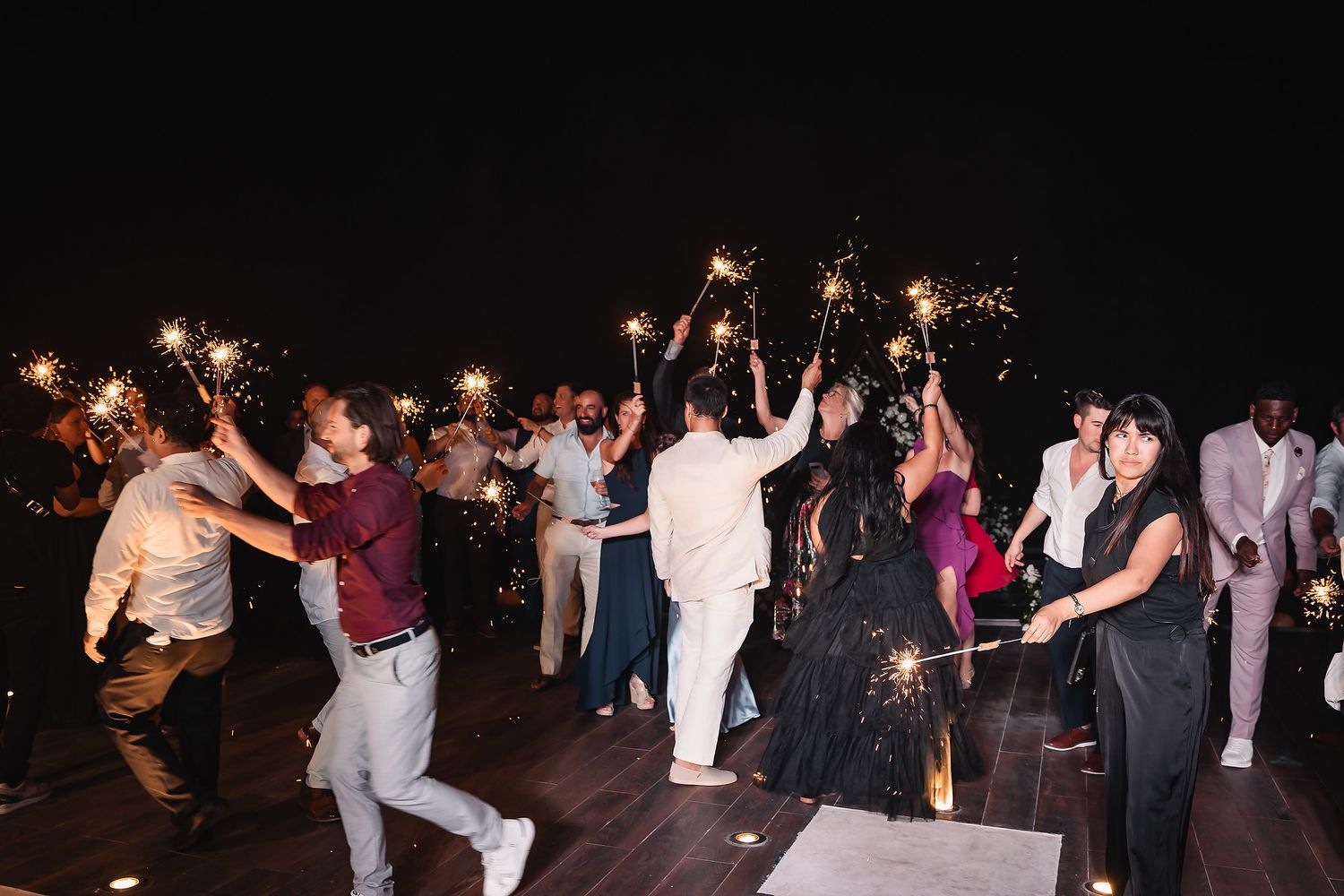 Wedding guests dance joyfully while holding sparklers during an outdoor nighttime celebration.
