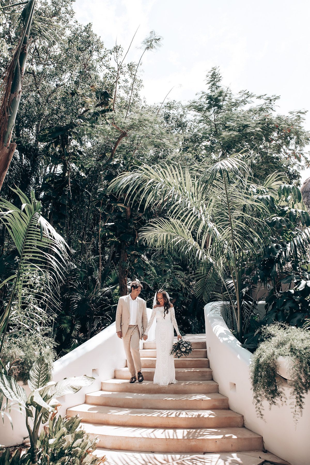 Wedding couple walking up white stone steps surrounded by lush tropical gardens and palm trees.