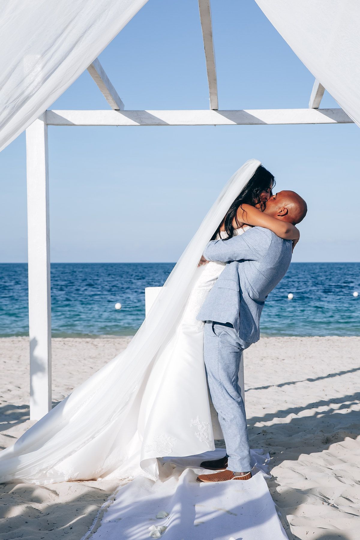 Romantic beach wedding photos showing couple embracing under flowing white fabric by the ocean.