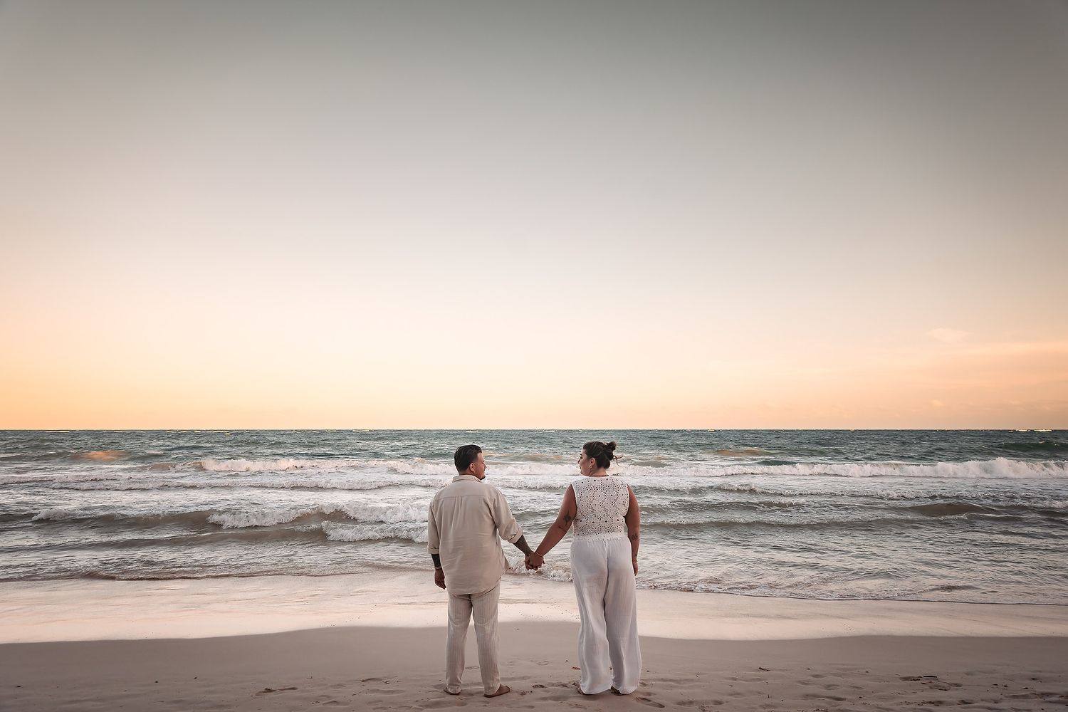 Couple in white attire walking along beach at sunset.