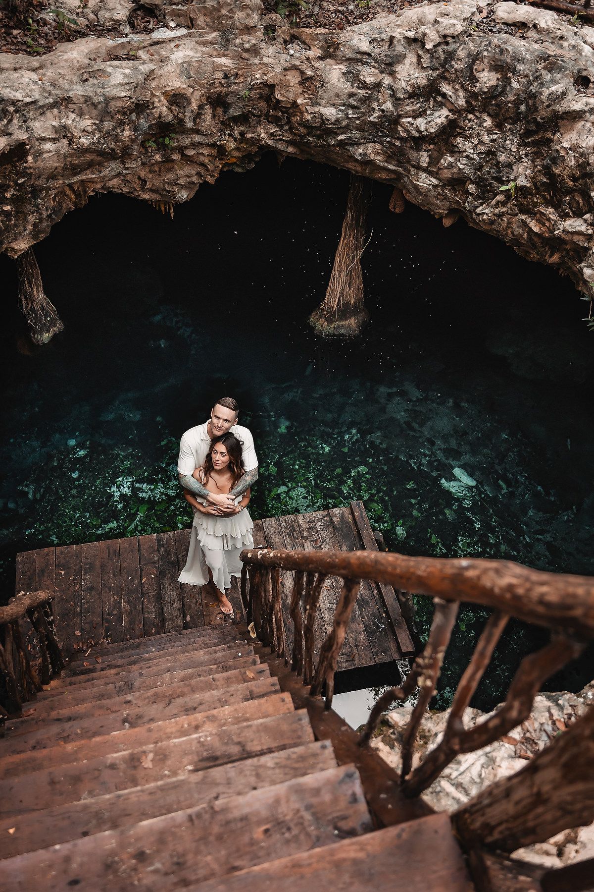 A couple stands on wooden stairs descending into a dark cave with turquoise water below.
