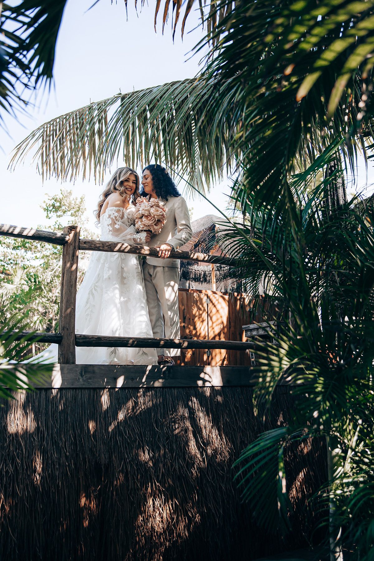 Couple poses together on a wooden deck surrounded by tropical greenery and palm trees.