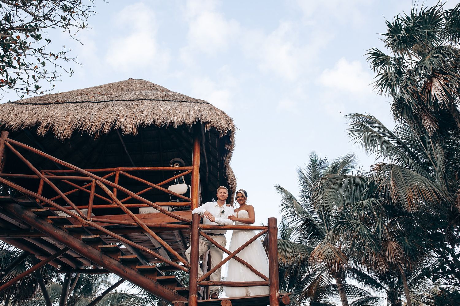 A couple poses on the balcony of a thatched-roof tropical beach hut surrounded by palm trees.