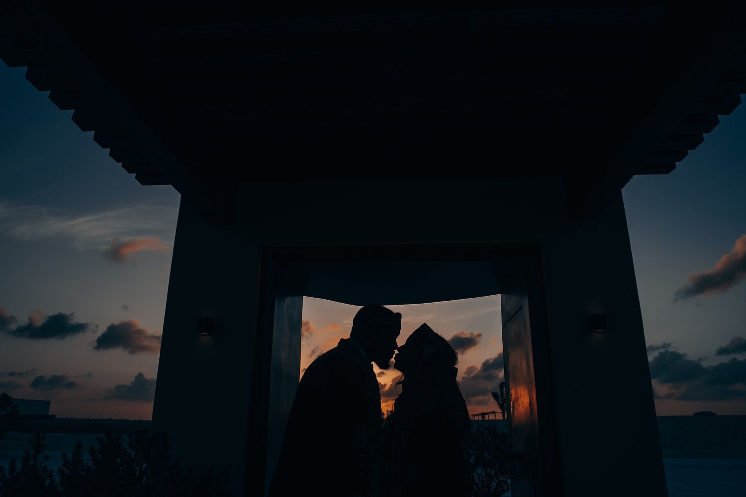 Silhouetted couple sharing intimate moment at sunset through architectural frame.