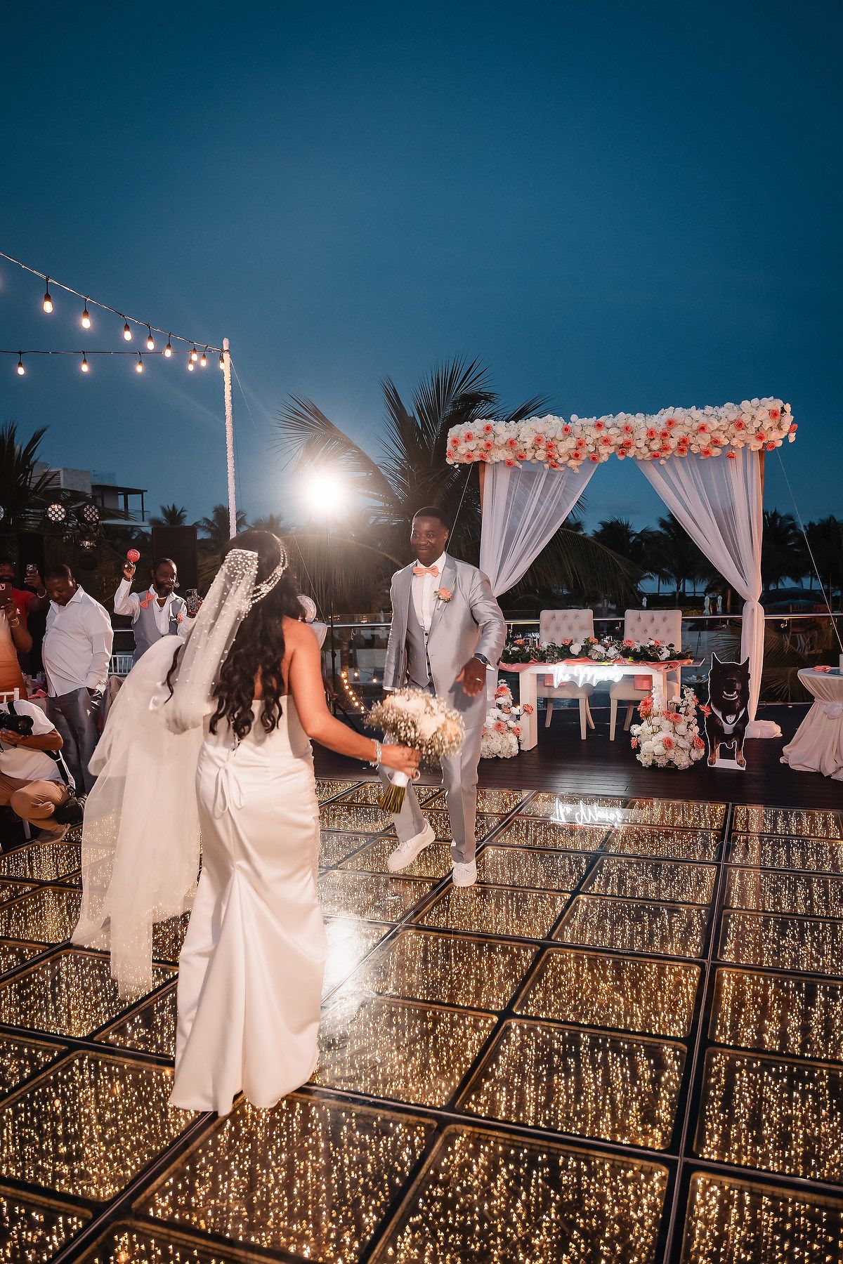 A romantic evening wedding reception with string lights and a floral arch on an illuminated dance floor.
