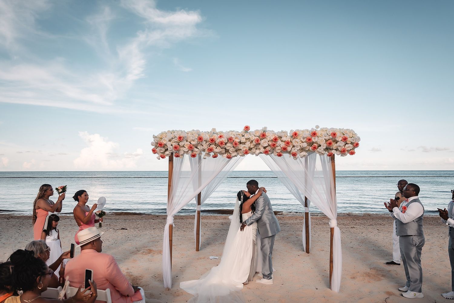 Wedding guests seated on a beach with a white draped arch and coral flowers for an oceanside ceremony.