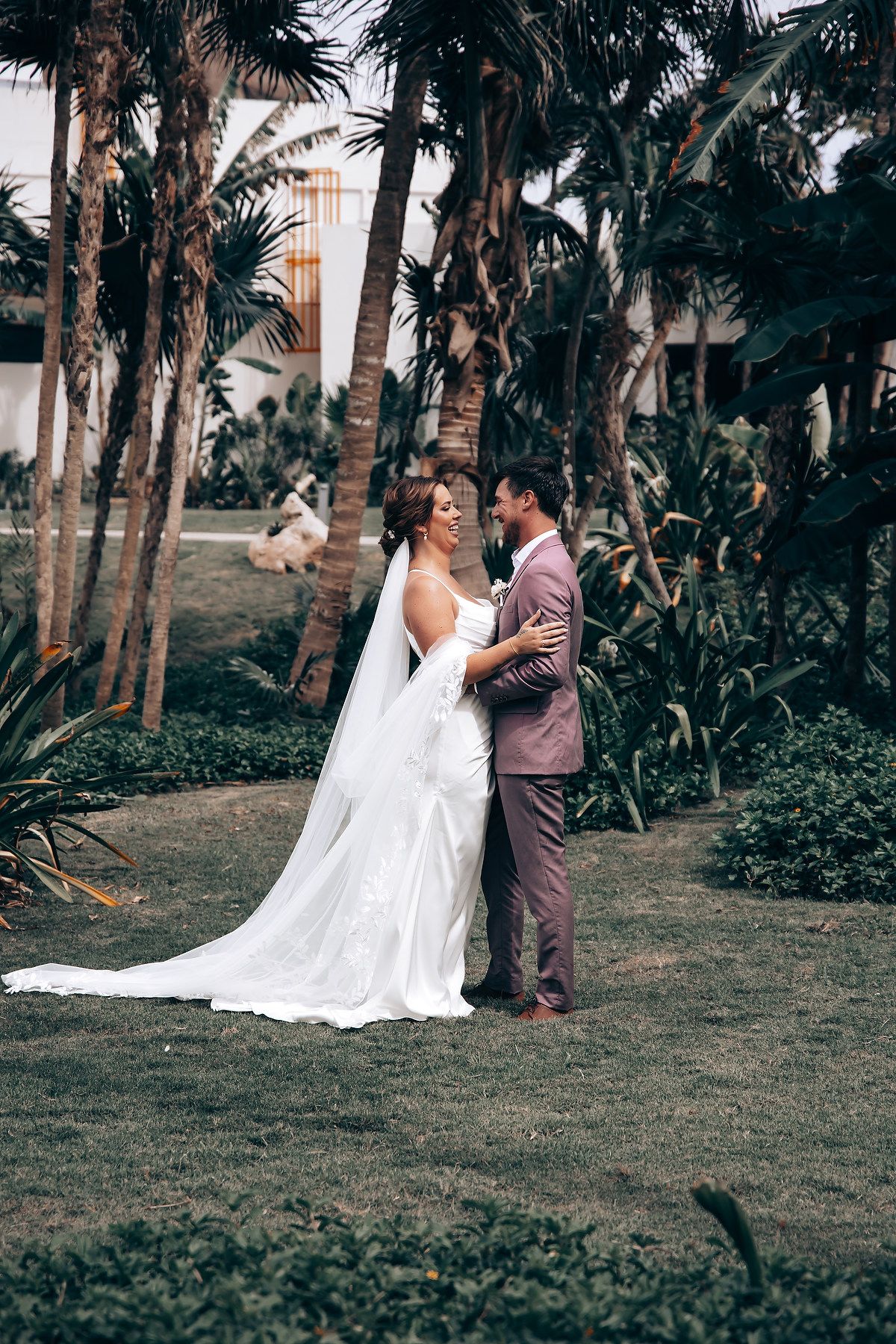 A couple shares a romantic moment among palm trees in a tropical garden setting during their wedding photoshoot.