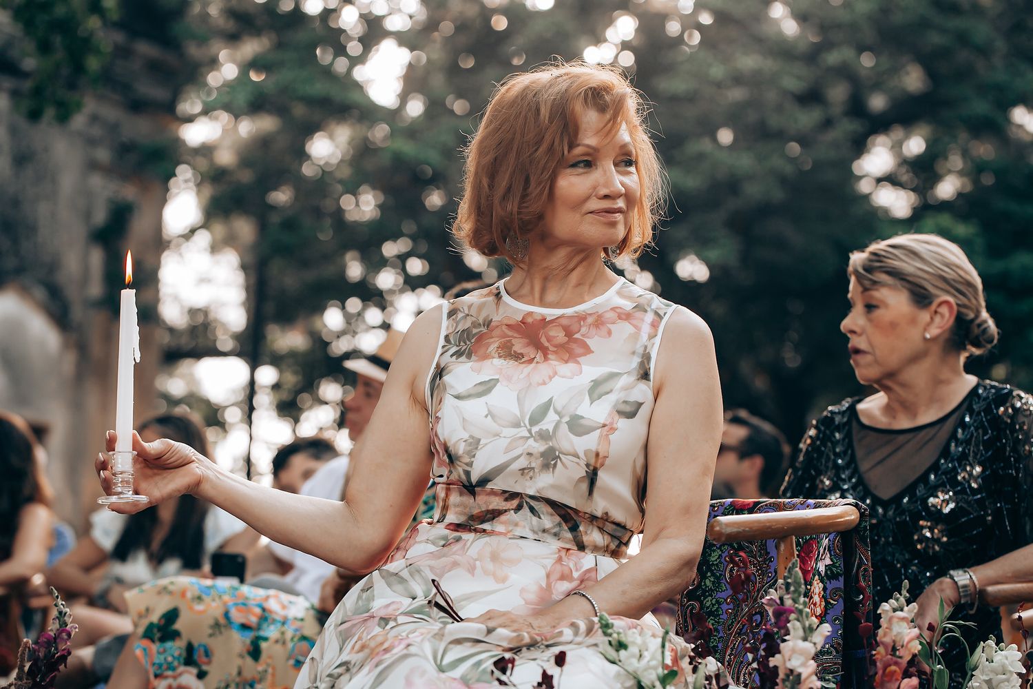 A woman in a floral dress sits outdoors at a summer gathering while another person is visible in the background.