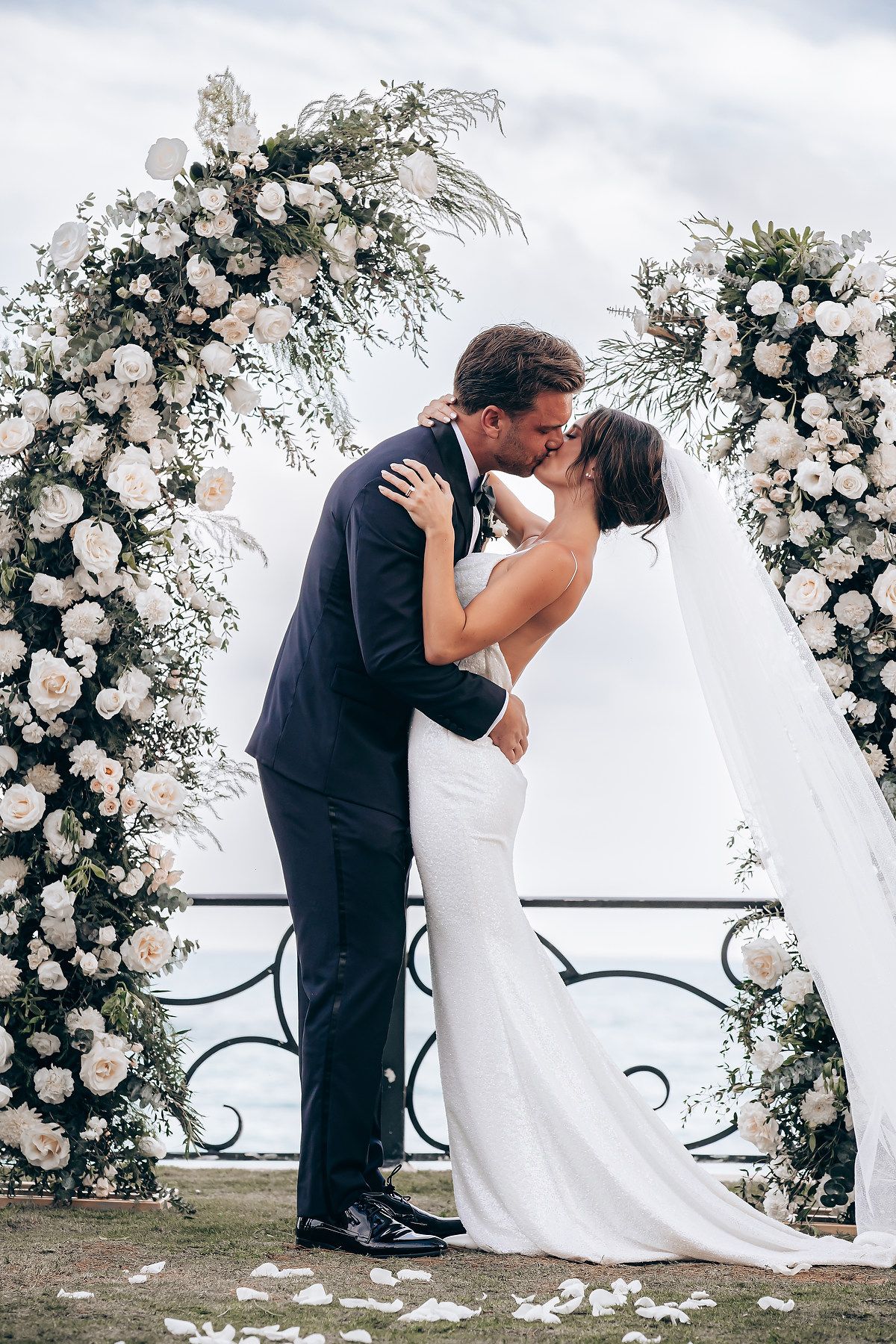 A romantic wedding ceremony under a beautiful floral arch with white roses and greenery against a cloudy sky.