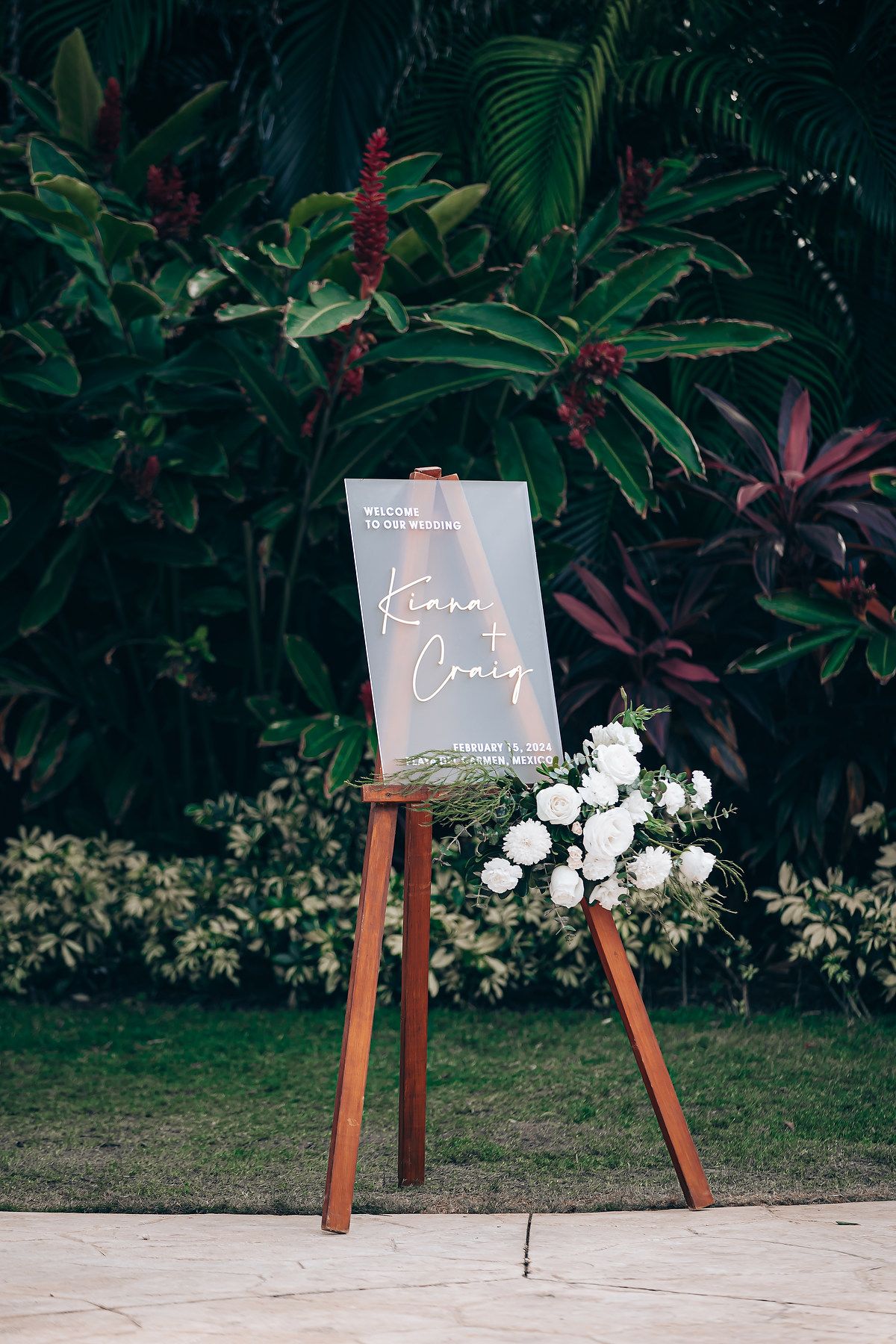 Modern acrylic wedding welcome signs displayed on wooden easels against lush tropical garden backdrop.