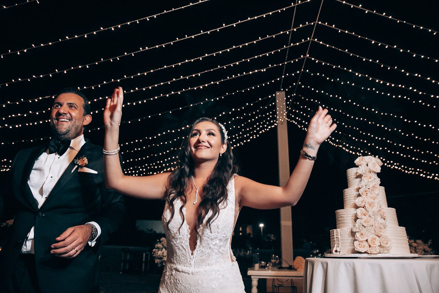 Wedding celebration under twinkling string lights with tiered cake in background.