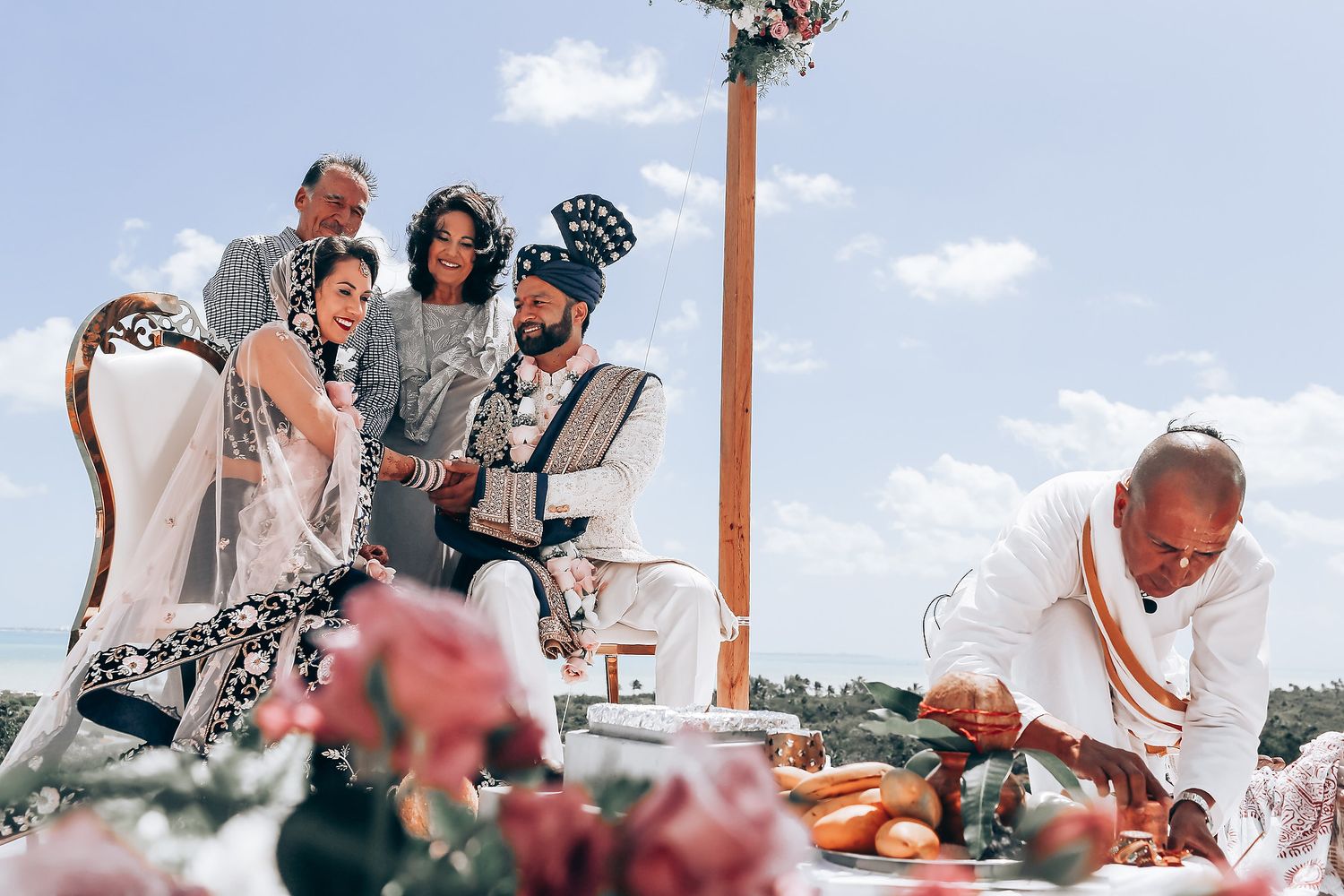 Wedding ceremony guests gathered outdoors under blue skies with wooden beams overhead.