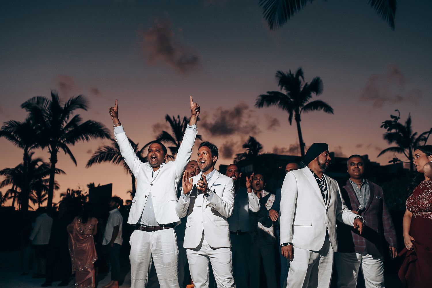 Wedding guests in white attire dance and celebrate at an outdoor reception during sunset with palm trees in background.