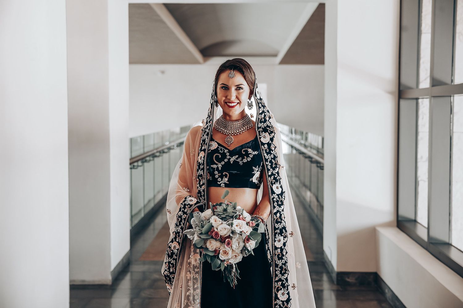 A person wearing a black and white floral dress stands confidently in a bright hallway with white columns.