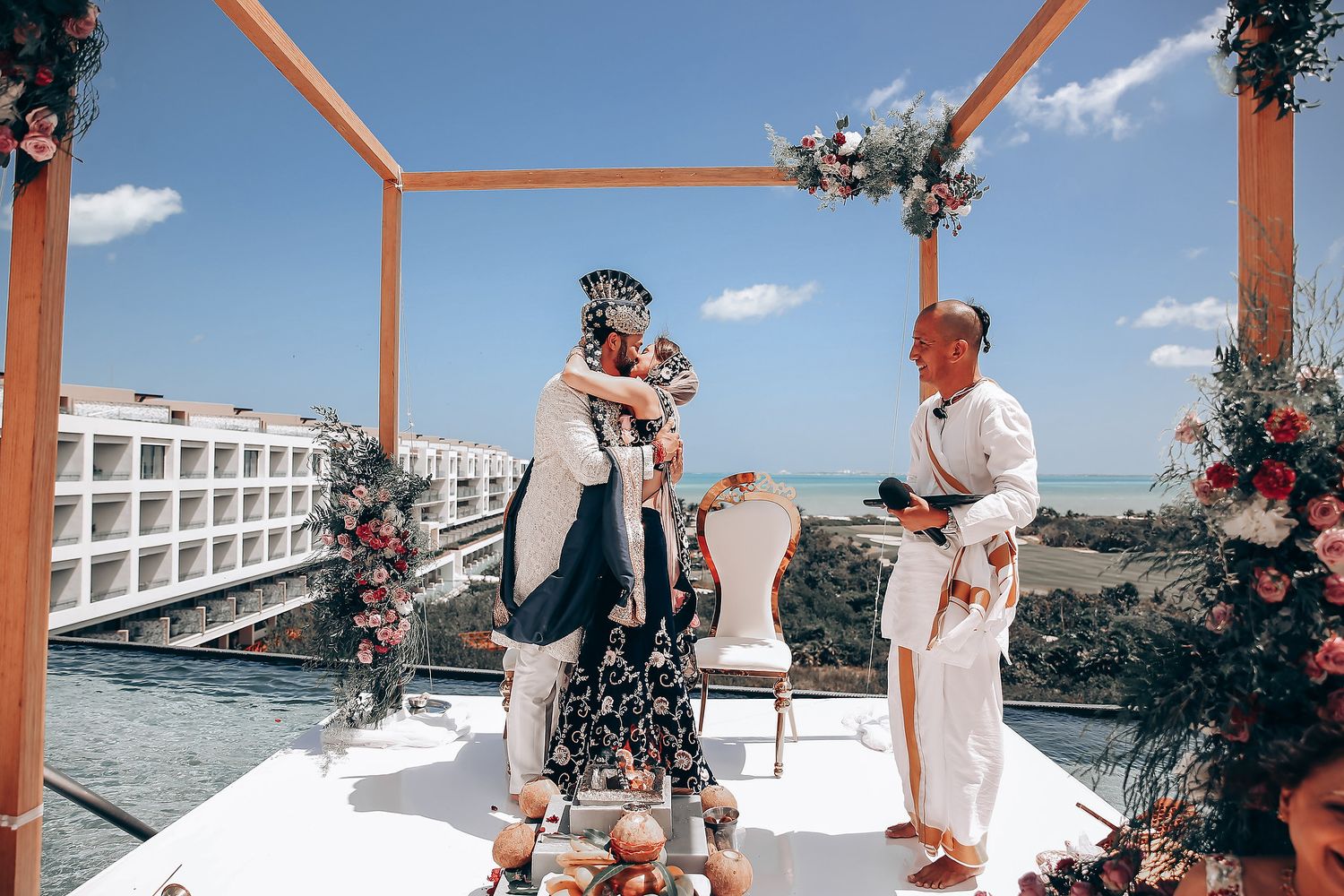 A wedding ceremony takes place under a coral-colored arch decorated with flowers on a sunny rooftop overlooking the ocean.