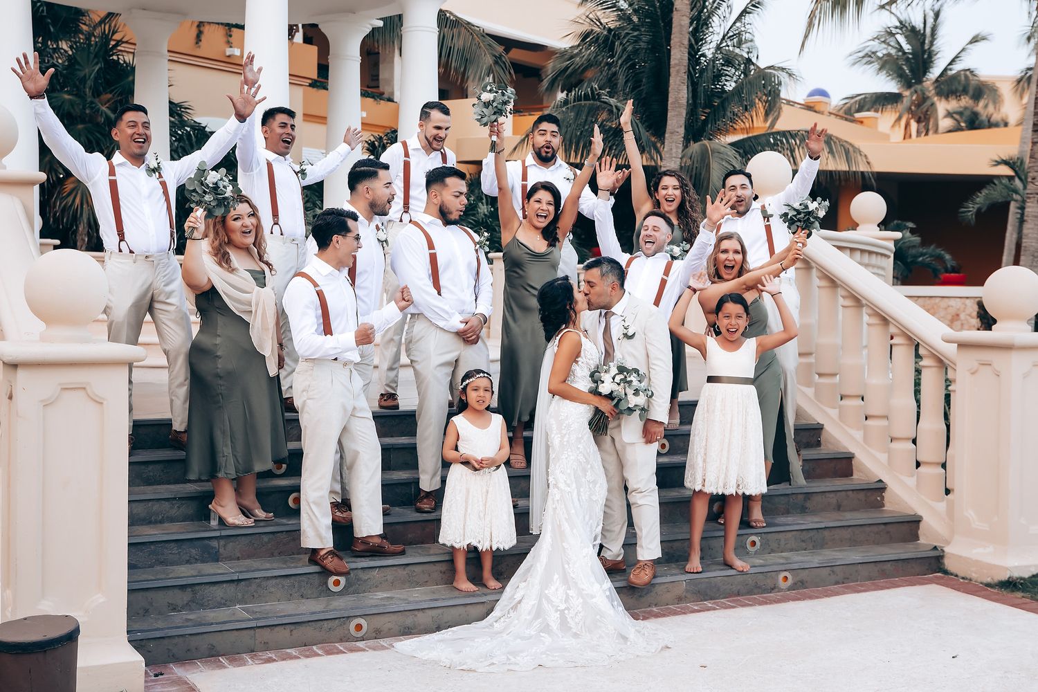 Wedding party dressed in white and red poses on elegant outdoor staircase at tropical venue during sunset celebration.