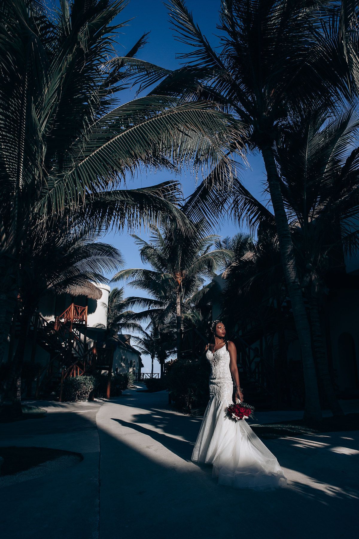 A person in a white wedding dress stands in dramatic lighting beneath swaying palm trees against a deep blue sky.