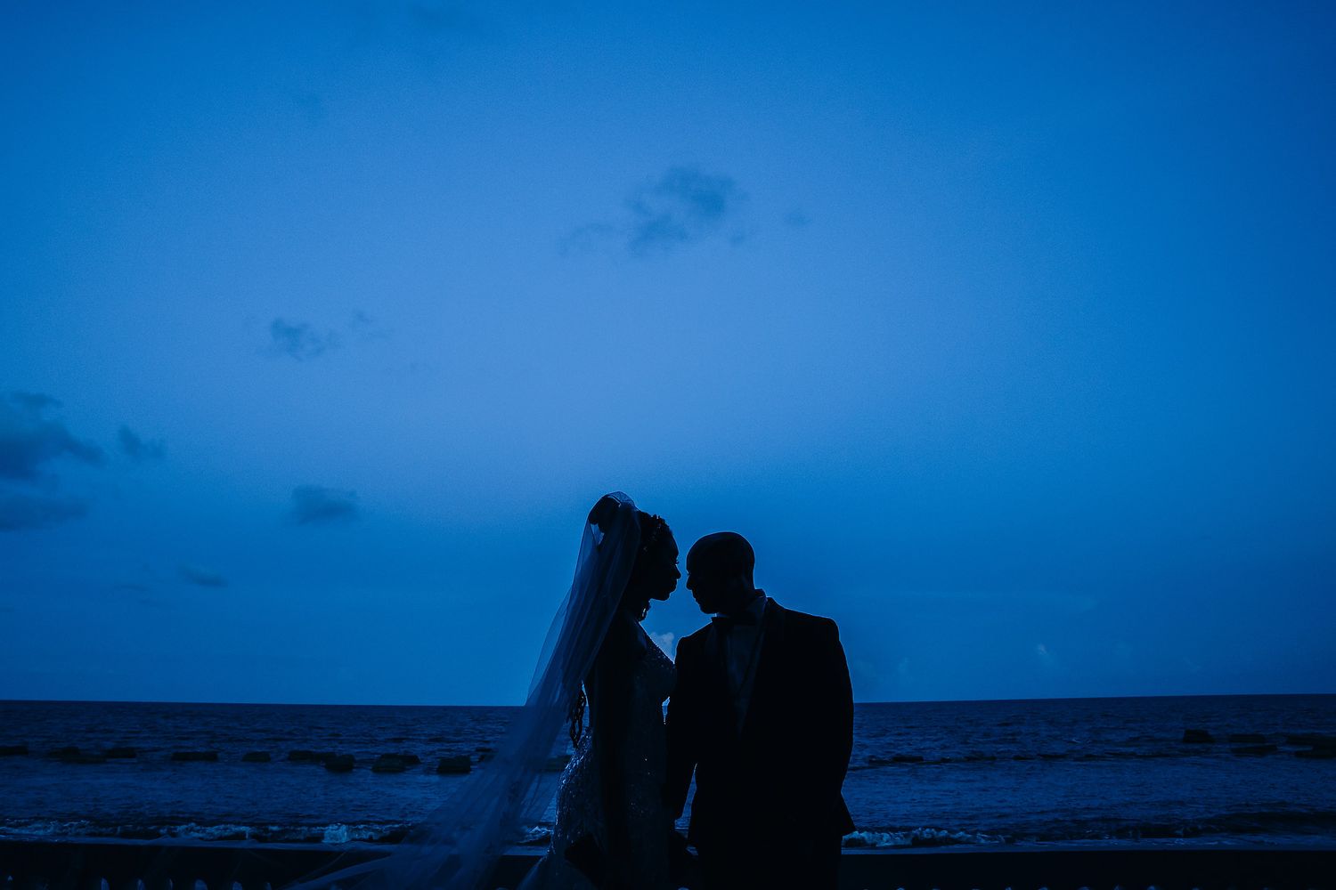 Silhouette of newlyweds standing by the ocean at dusk against a deep blue sky.