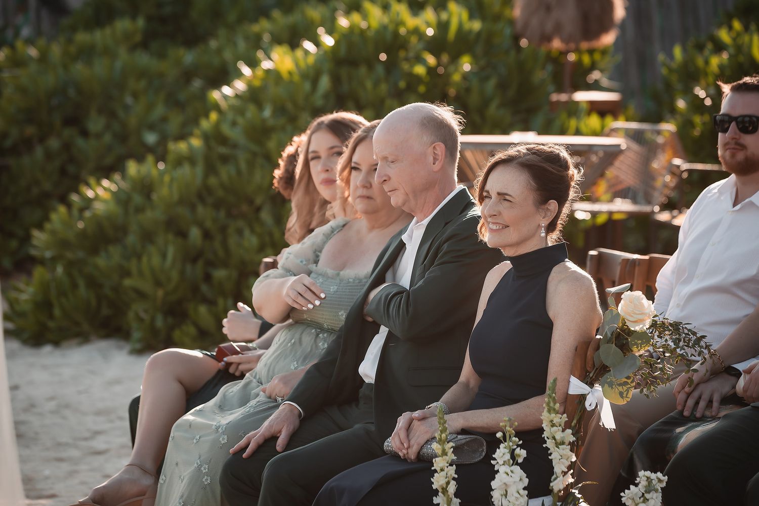 Guests smiling and seated outdoors at a beach wedding ceremony, dressed in elegant attire on a sunny day.